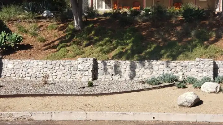 A long river rock retaining wall holding a planted slope above a gravel yard